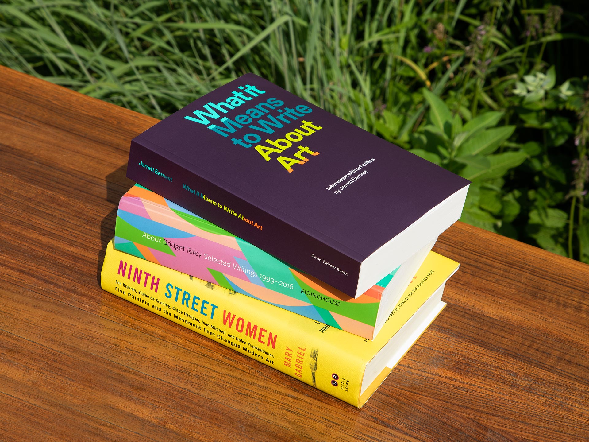 A photograph of three books stacked on a bench outside. The titles are: What it Means to Write About Art: Interviews with art critics; About Bridget Riley: Selected Writings 1999–2016; Ninth Street Women: Lee Krasner, Elaine De Kooning, Grace.